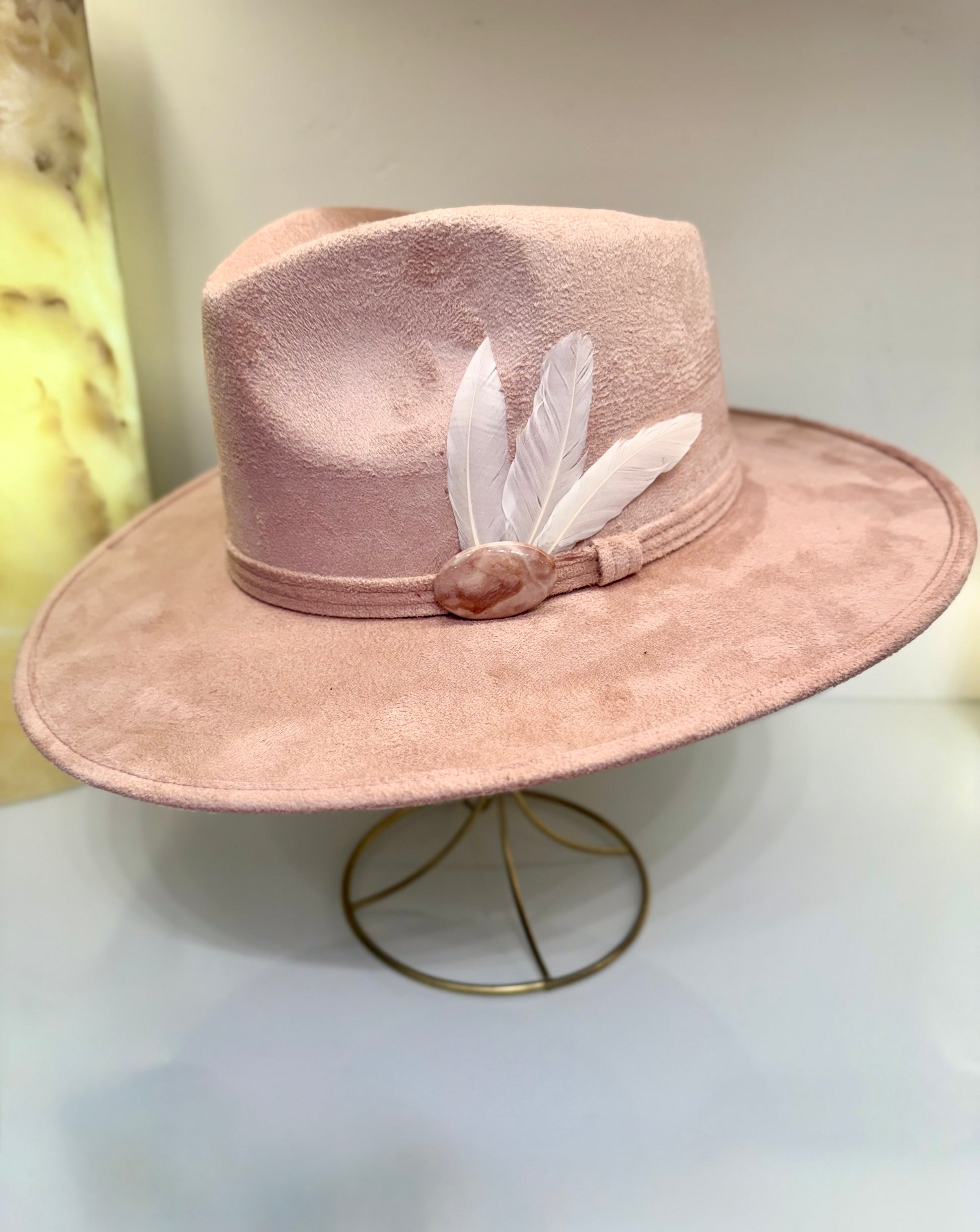 A dusty pink felt wide-brim hat with a matching band, decorated with two white feathers and a small pink shell accent, displayed on a gold wire stand.